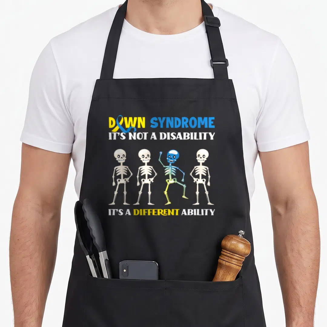 Month down syndrome awareness apron - Four cartoon skeletons, one rainbow-colored waving, with awareness text and ribbon — shown on a black cotton canvas apron against a white background