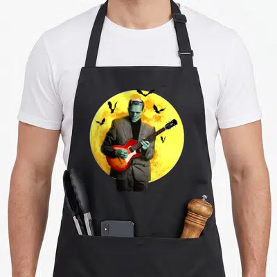 Frankenguitar halloween apron - Frankenstein in grey suit plays red guitar before yellow moon with black bats — shown on a black cotton canvas apron against a white background