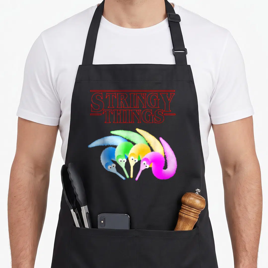 Stringy magic apron - Red outline text reading Stringy Things above four fuzzy worms in blue, green, yellow, and pink — shown on a black cotton canvas apron against a white background