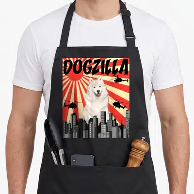 Japanese samoyed apron - DOGZILLA text above giant white dog behind city skyline with flying helicopters on red — shown on a black cotton canvas apron against a white background