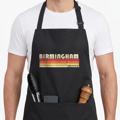 Root alabama apron - BIRMINGHAM text above red orange yellow cream stripes with ALABAMA on bottom right — shown on a black cotton canvas apron against a white background