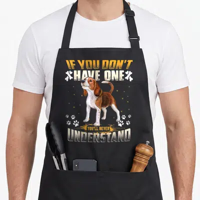 Youll dog apron - Beagle standing between orange and white text flanked by bones and scattered paw prints — shown on a black cotton canvas apron against a white background