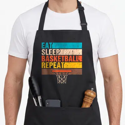 Sleep game over thirty apron - EAT SLEEP BASKETBALL REPEAT text in colored blocks above a basketball hoop net — shown on a black cotton canvas apron against a white background
