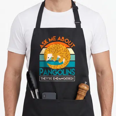 Save im hoot apron - Retro orange pangolin over striped sun background with endangered awareness text — shown on a black cotton canvas apron against a white background