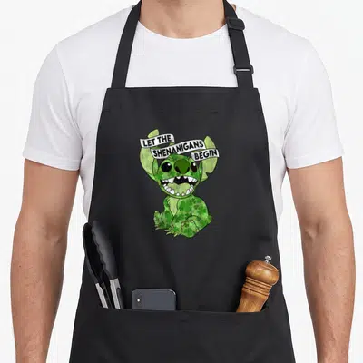 Funny stitch st patrick day aprons - Green Stitch covered in shamrocks with white banner reading LET THE SHENANIGANS — shown on a black cotton canvas apron against a white background
