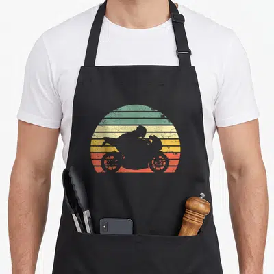 Silhouette biker apron - Grey motorcyclist silhouette against striped sun in green yellow orange red grey retro style — shown on a black cotton canvas apron against a white background