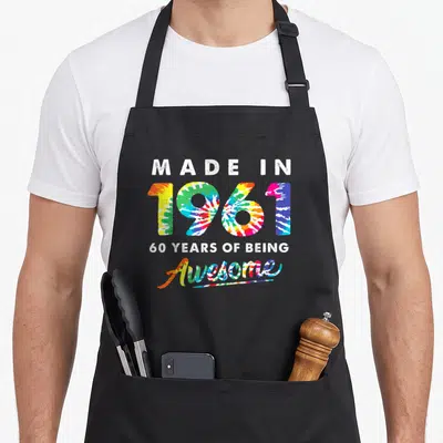 60th tie apron - Stacked text Made in 1961 60 Years Of Being Awesome in tie-dye colors — shown on a black cotton canvas apron against a white background