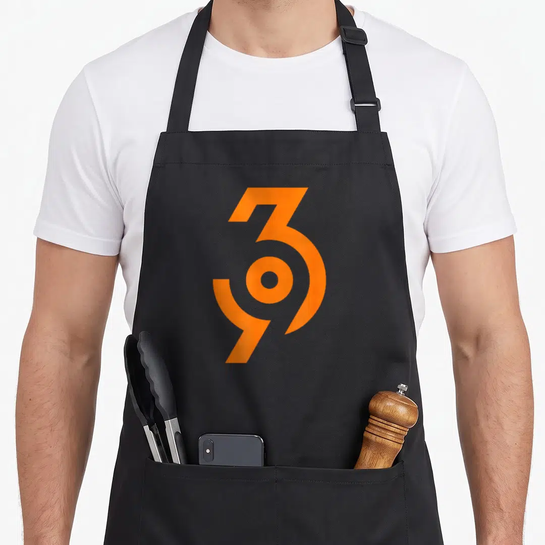 Method manifestation apron - Orange abstract geometric symbol centered on grey background — shown on a black cotton canvas apron against a white background