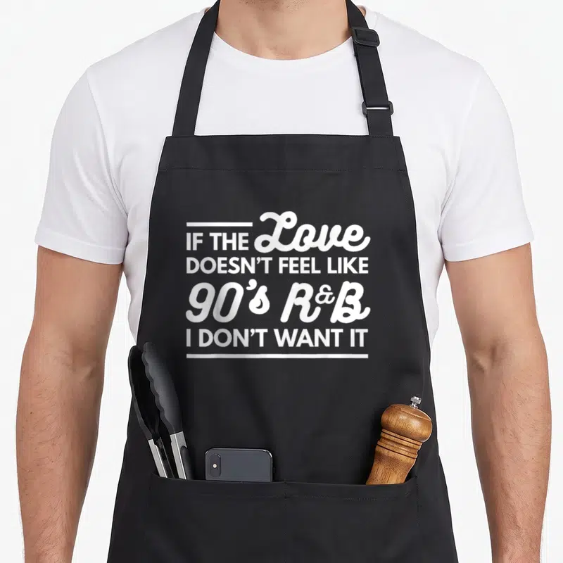 Nostalgia r aprons - White stacked text reading IF THE Love DOESN'T FEEL LIKE 90's R&B I DON'T WANT IT framed by two — shown on a black cotton canvas apron against a white background