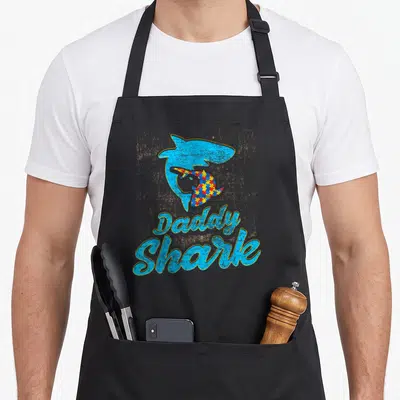 Autism daddy apron - Blue shark with rainbow puzzle pattern and Daddy Shark text — shown on a black cotton canvas apron against a white background