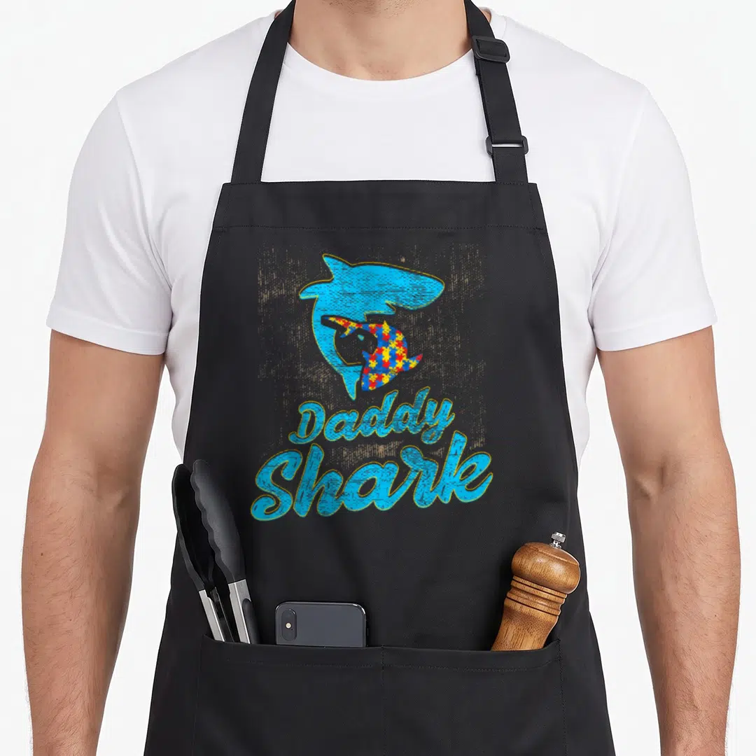 Autism daddy apron - Blue shark with rainbow puzzle pattern and Daddy Shark text — shown on a black cotton canvas apron against a white background