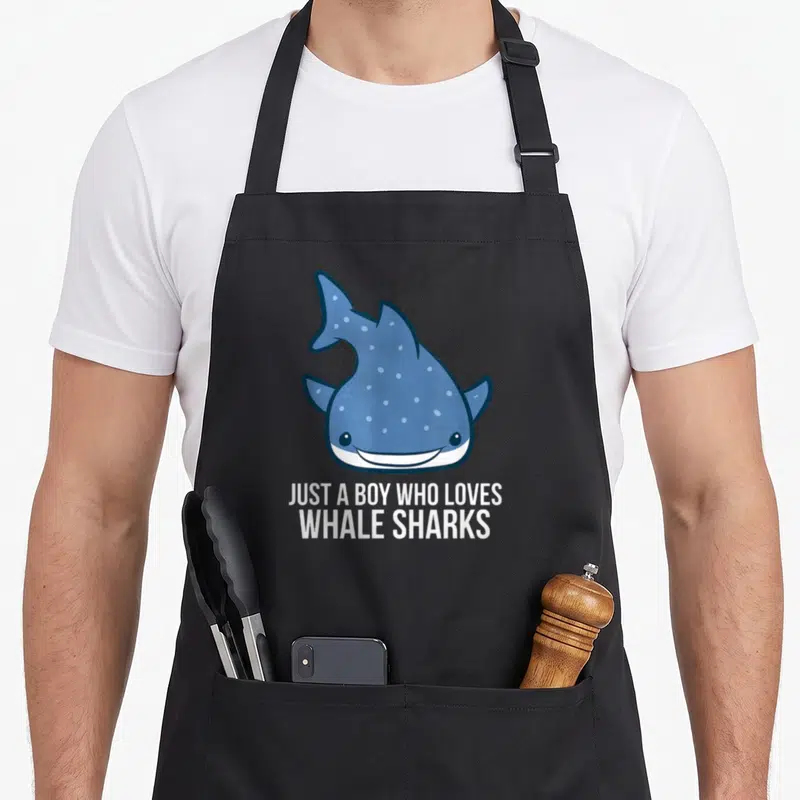 Cute whale shark apron - Blue cartoon whale shark above white text reading JUST A BOY WHO LOVES WHALE SHARKS — shown on a black cotton canvas apron against a white background