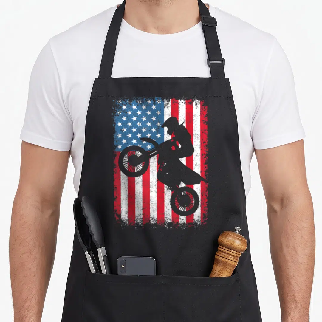Dirt 4th of july apron - Grey motocross rider silhouette on distressed red white blue American flag background — shown on a black cotton canvas apron against a white background