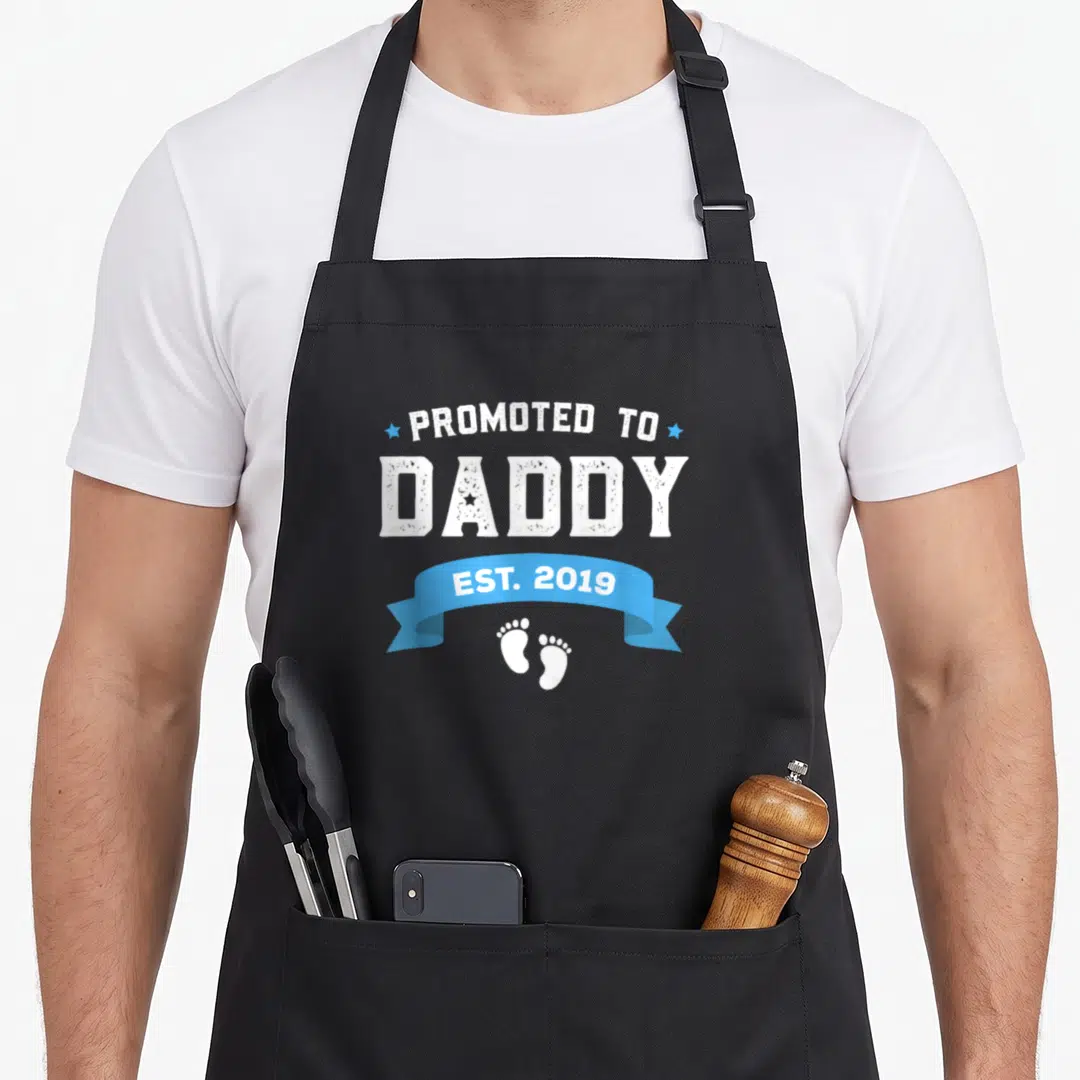 Vintage promoted daddy apron - PROMOTED TO DADDY EST 2019 text with blue ribbon and white baby footprints in distressed — shown on a black cotton canvas apron against a white background