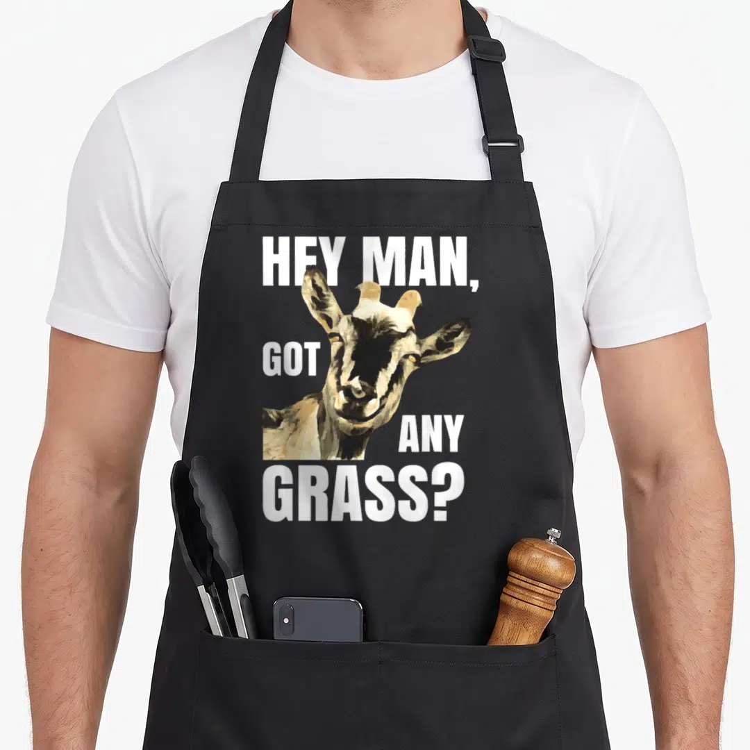 Animal farm apron - Cartoon goat head with white text hey man got any grass wrapped around it — shown on a black cotton canvas apron against a white background