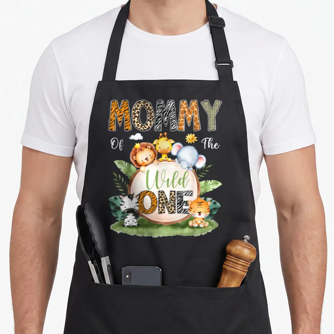 1st safari birthday apron - MOMMY Of The Wild ONE text with cartoon animals and peach circle — shown on a black cotton canvas apron against a white background