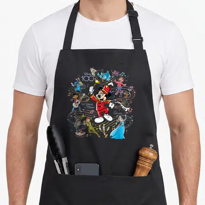 Music novelty apron - Mickey Mouse conductor surrounded by Disney characters playing instruments with 100 Years — shown on a black cotton canvas apron against a white background