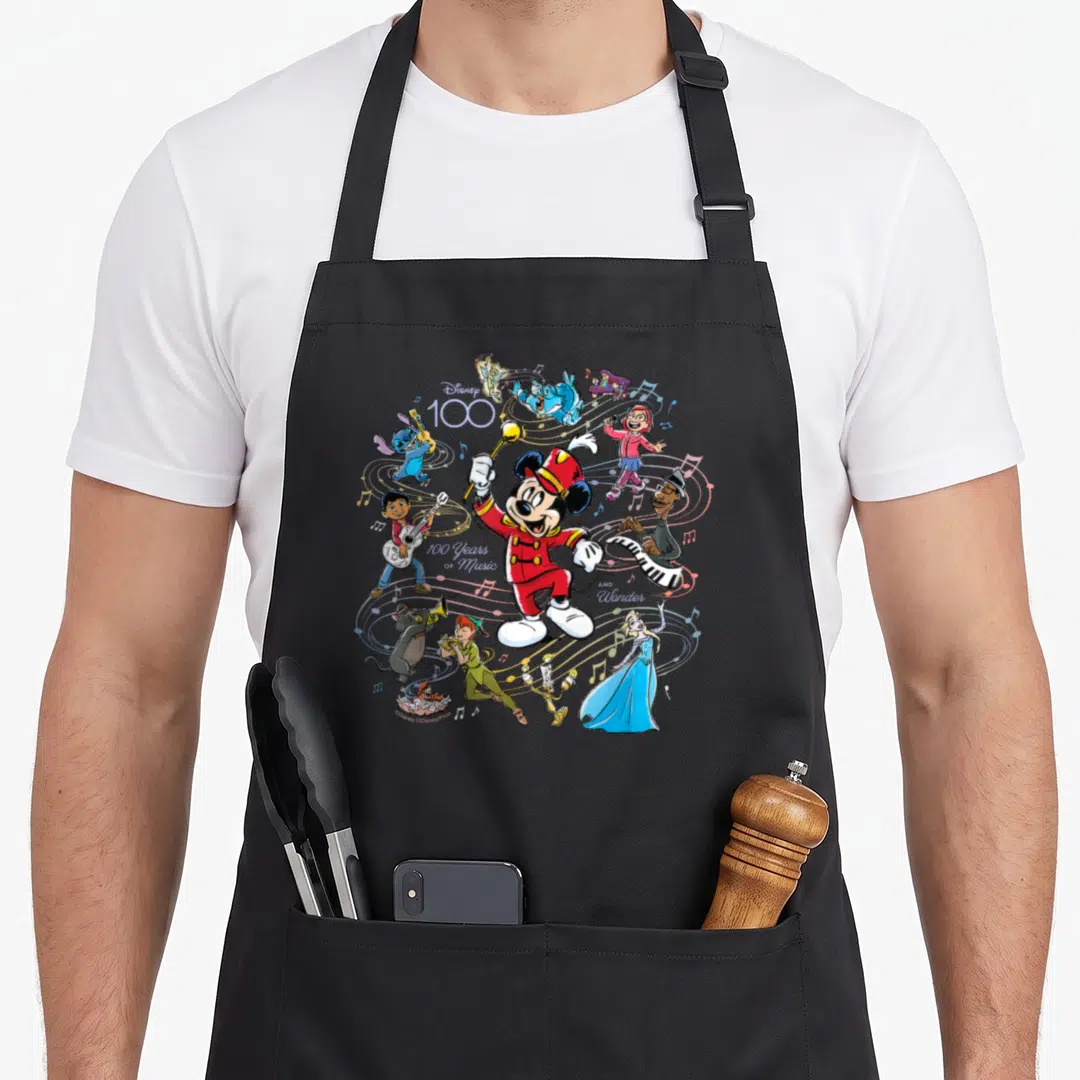 Music novelty apron - Mickey Mouse conductor surrounded by Disney characters playing instruments with 100 Years — shown on a black cotton canvas apron against a white background