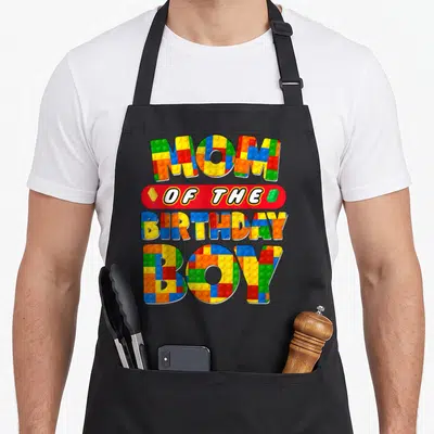 Decor building aprons - Colorful brick-textured text reading Mom of the Birthday Boy stacked on four lines — shown on a black cotton canvas apron against a white background