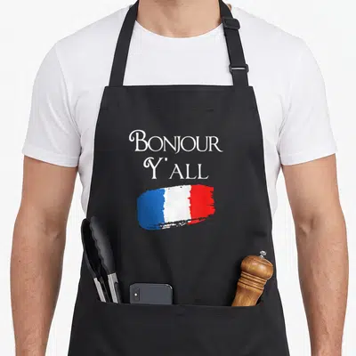 Bonjour french apron - BONJOUR Y'ALL text above blue white red brush-stroke French flag on gray background — shown on a black cotton canvas apron against a white background