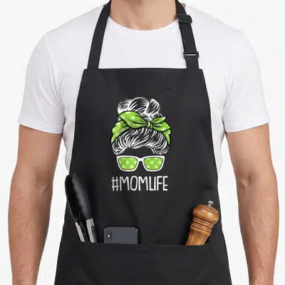 Weed mother's day aprons - White cartoon head with green leaf bandana and sunglasses above #MOMLIFE text — shown on a black cotton canvas apron against a white background