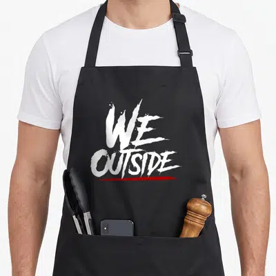 Outside novelty apron - White We Outside text with red brush underline on grey background — shown on a black cotton canvas apron against a white background
