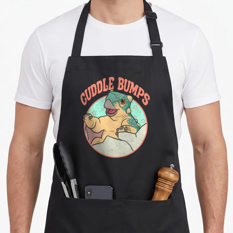 Jurassic lover comfort apron - Cartoon dinosaur reaching from teal circle under arched CUDDLE BUMPS text in coral — shown on a black cotton canvas apron against a white background