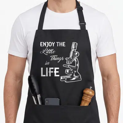 Microbiologist science apron - White line art microscope next to stacked text ENJOY THE Little Things in LIFE — shown on a black cotton canvas apron against a white background