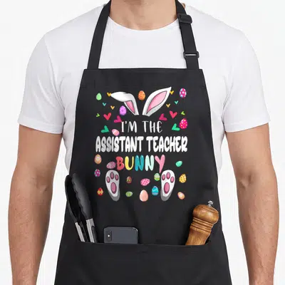 Assistant easter bunny aprons - Cartoon bunny ears paws text I'M THE ASSISTANT TEACHER BUNNY with colorful eggs — shown on a black cotton canvas apron against a white background