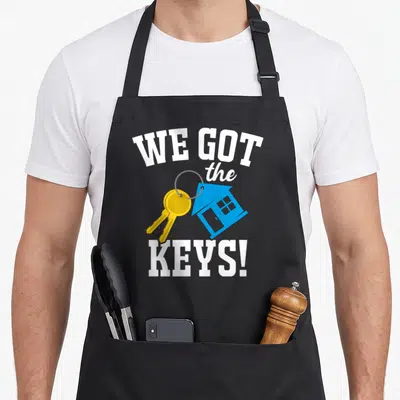 Bought homeowner novelty apron - Cartoon keys and blue house icon with WE GOT the KEYS! text — shown on a black cotton canvas apron against a white background