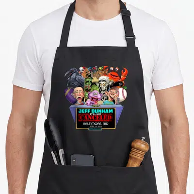 Dunham hollywood apron - Ventriloquist puppets on blue stage with JEFF DUNHAM STILL NOT CANCELED BALTIMORE text — shown on a black cotton canvas apron against a white background