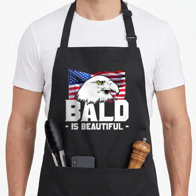 Beautiful eagles apron - Realistic bald eagle head over waving US flag with white text reading BALD IS BEAUTIFUL — shown on a black cotton canvas apron against a white background