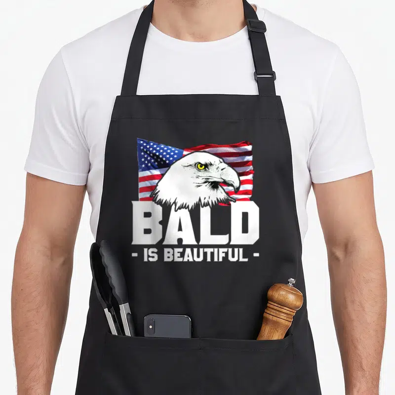 Beautiful eagles apron - Realistic bald eagle head over waving US flag with white text reading BALD IS BEAUTIFUL — shown on a black cotton canvas apron against a white background