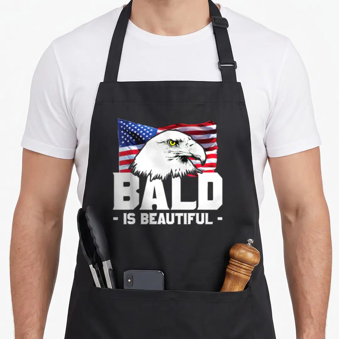 Beautiful eagles apron - Realistic bald eagle head over waving US flag with white text reading BALD IS BEAUTIFUL — shown on a black cotton canvas apron against a white background