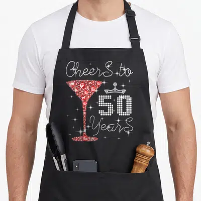 Princess queen apron - Rhinestone cocktail glass and crown with Cheers To 50 Years text — shown on a black cotton canvas apron against a white background