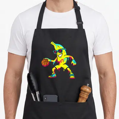 Basketball banana apron - Cartoon yellow banana character in blue sunglasses and shorts holding an orange basketball — shown on a black cotton canvas apron against a white background