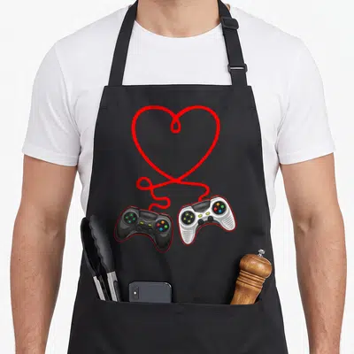 Gamer valentine apron - Two game controllers connected by a red heart-shaped cable on black background — shown on a black cotton canvas apron against a white background