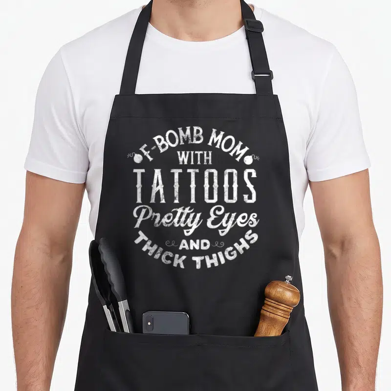 Bomb tattoo apron - White distressed text F-BOMB MOM WITH TATTOOS Pretty Eyes AND THICK THIGHS with two small bombs — shown on a black cotton canvas apron against a white background