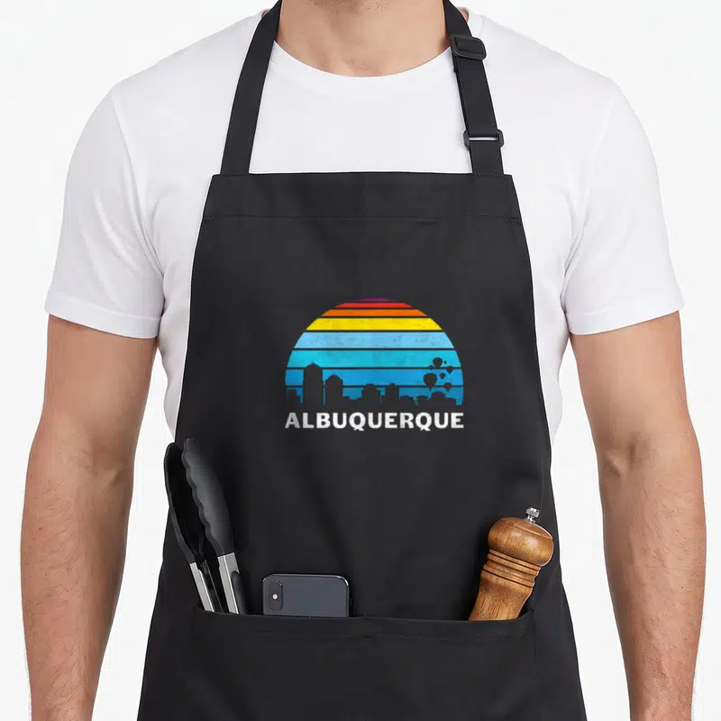Downtown albuquerque apron - Retro blue skyline with orange and yellow striped sun and white Albuquerque text — shown on a black cotton canvas apron against a white background