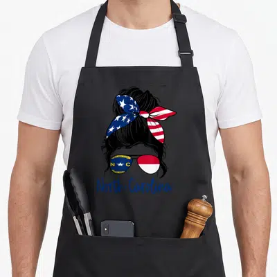 North messy aprons - Woman silhouette with messy bun, flag sunglasses, and North Carolina text in blue red white — shown on a black cotton canvas apron against a white background