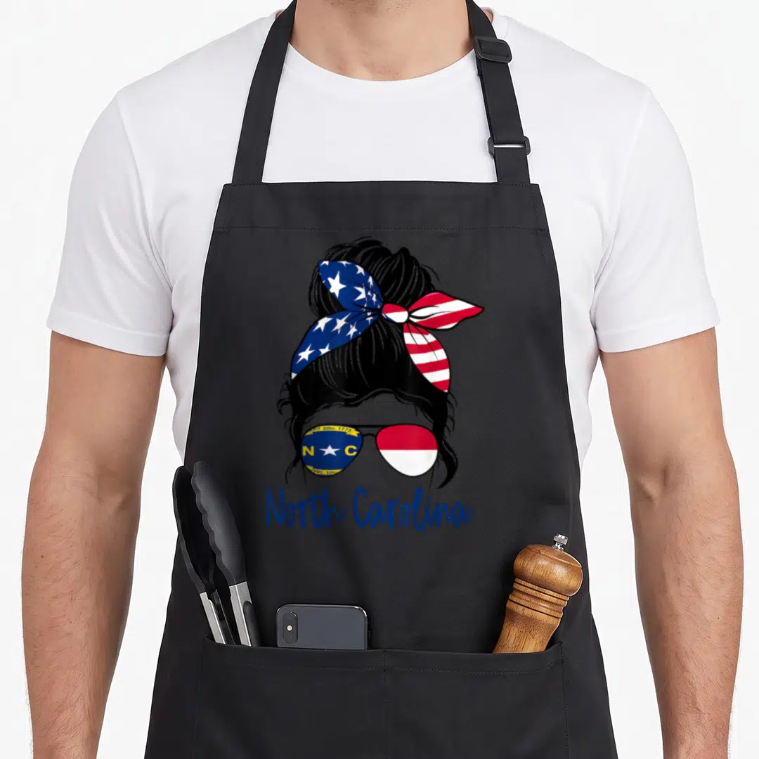 North messy aprons - Woman silhouette with messy bun, flag sunglasses, and North Carolina text in blue red white — shown on a black cotton canvas apron against a white background
