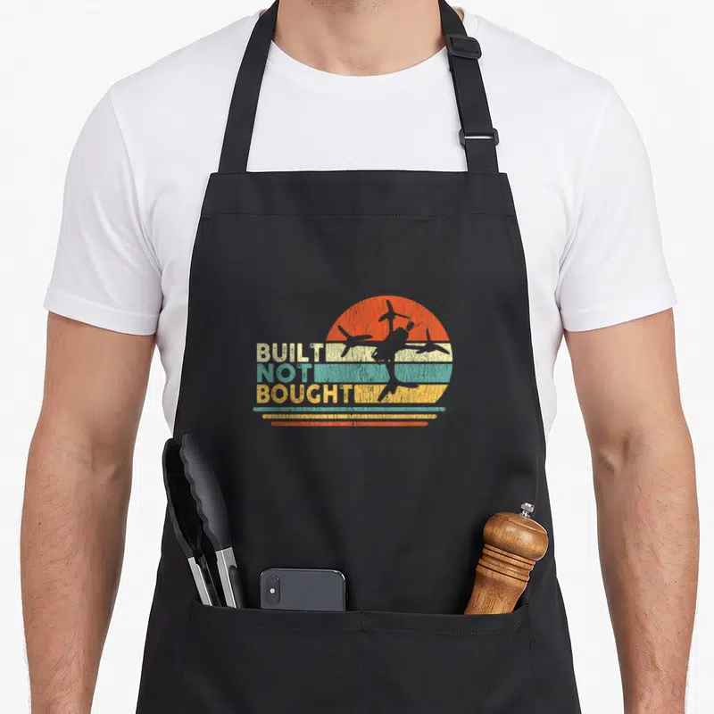 Built builder apron - BUILT NOT BOUGHT text with retro airplane silhouette on striped orange teal yellow beige sun — shown on a black cotton canvas apron against a white background