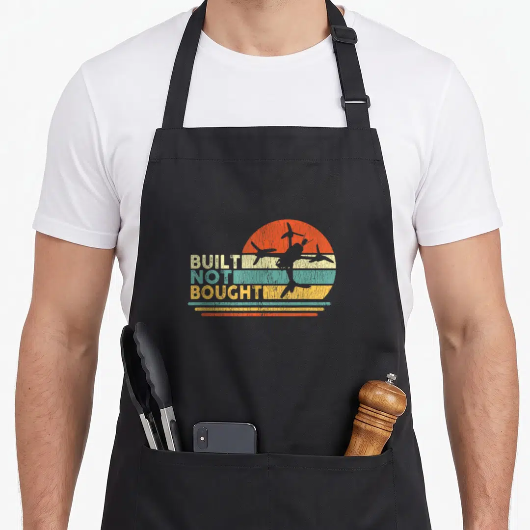 Built builder apron - BUILT NOT BOUGHT text with retro airplane silhouette on striped orange teal yellow beige sun — shown on a black cotton canvas apron against a white background