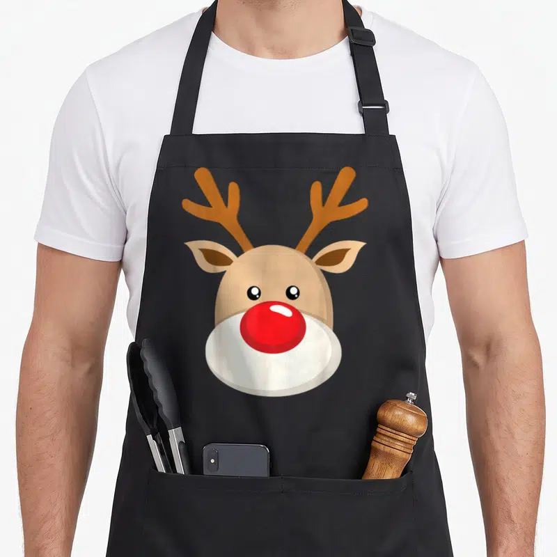 Antler red christmas apron - Cartoon reindeer head with large red nose, tan antlers, and white muzzle — shown on a black cotton canvas apron against a white background