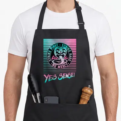 Cobra kai ken apron - Retro cobra logo with strike first text over pink blue stripes and yes sensei below — shown on a black cotton canvas apron against a white background