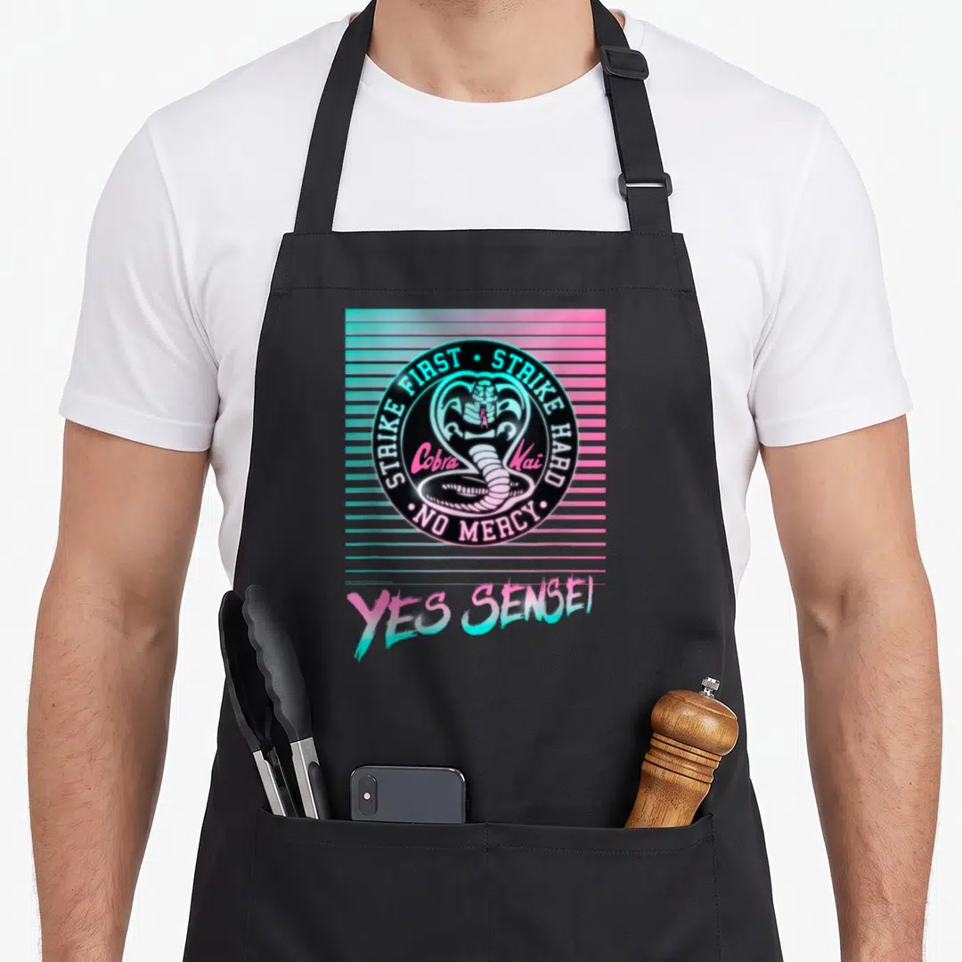 Cobra kai ken apron - Retro cobra logo with strike first text over pink blue stripes and yes sensei below — shown on a black cotton canvas apron against a white background