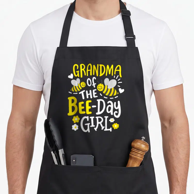 Grandma birthday bee aprons - Stacked yellow and white text GRANDMA OF THE BEE-DAY GIRL with two smiling bees — shown on a black cotton canvas apron against a white background