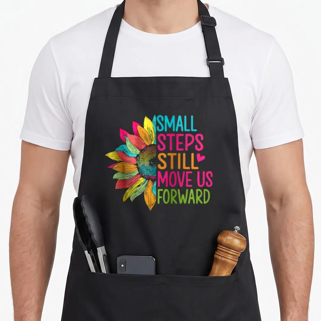 Move us aprons - Half sunflower with pink yellow blue green orange petals beside stacked motivational text — shown on a black cotton canvas apron against a white background
