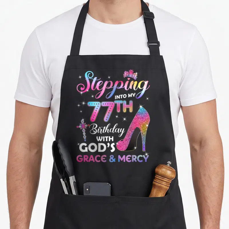 Stepping 77th birthday apron - Glittery high heel shoe next to Stepping Into My 77th Birthday With Gods Grace Mercy text — shown on a black cotton canvas apron against a white background