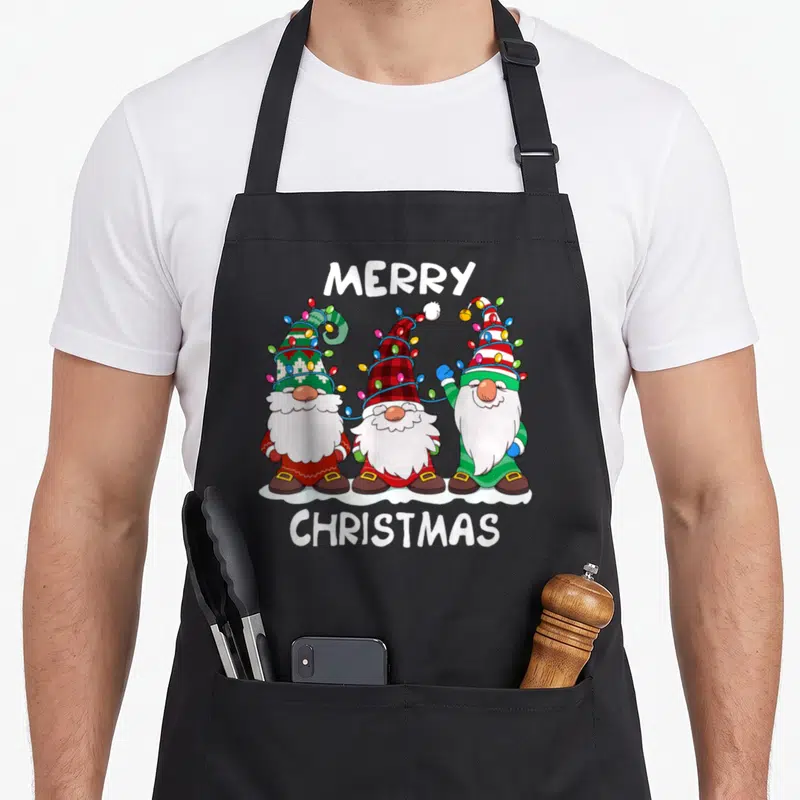 Gnome merry christmas apron - Three cartoon gnomes with beards wrapped in string lights under MERRY CHRISTMAS text — shown on a black cotton canvas apron against a white background